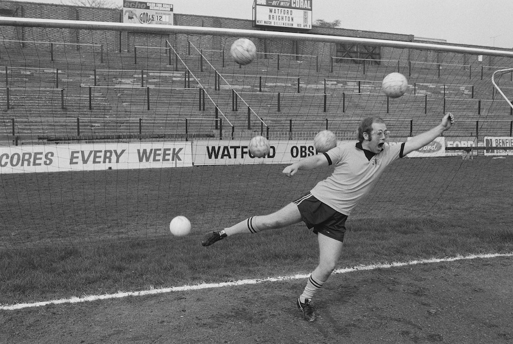 English singer-songwriter, and vice-president of Watford FC, Elton John, is overwhelmed by multiple shots on goal at the Vicarage Road stadium in Watford, April 1974. (Photo by Michael Putland/Getty Images)